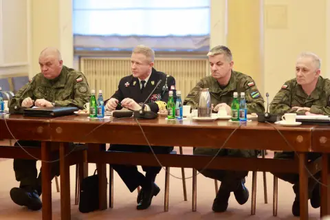 Czterech mężczyzn w mundurach siedzi przy drewnianym stole, przed nimi butelki wody i filiżanki.