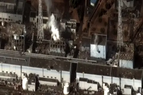 Widok z lotu ptaka na zniszczoną elektrownię Fukushima. Widać dym wydobywający się z uszkodzonych budynków, świadectwo katastrofy.