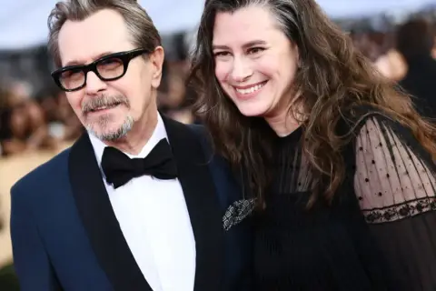Gary Oldman z żoną, oboje uśmiechnięci, on w eleganckim garniturze, ona w czarnej sukni z koronkowymi rękawami.