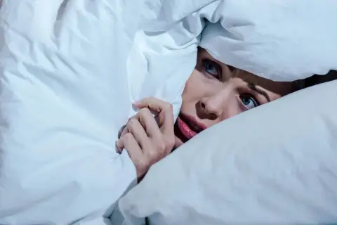 Verängstigte Frau versteckt sich unter der Decke, die Augen weit aufgerissen, als ob sie einen Albtraum erlebt.