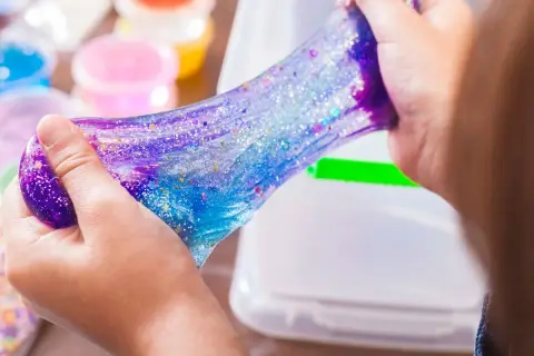 Dziecko bawi się błyszczącym, galaktycznym slime. W tle widać pojemniki z kolorowymi składnikami do slime, przepis na zabawę.