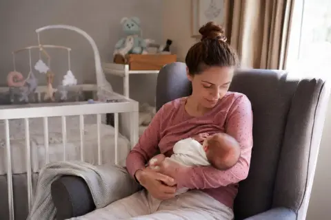 Matka karmi piersią noworodka w fotelu. W tle widać łóżeczko dziecięce i pluszowego misia. To piękny obrazek z okresu po porodzie.