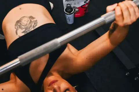 Mujer haciendo press de banca con barra. A su lado, un bote de proteína con leucina, esencial para la recuperación muscular.
