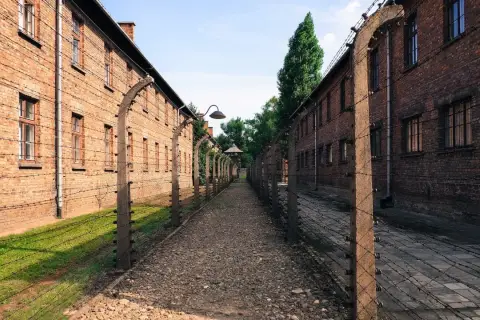 Ścieżka między ceglanymi budynkami, otoczona drutem kolczastym, prowadzi do wieży strażniczej. Miejsce pamięci Auschwitz Birkenau Museum.