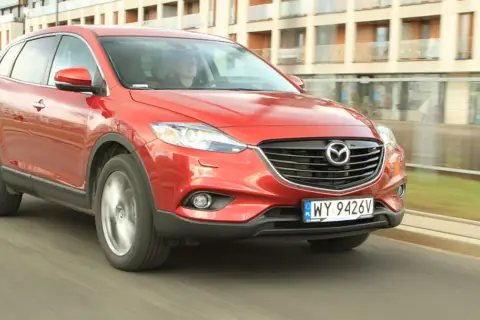 Czerwona Mazda CX-9 w ruchu miejskim, z widocznym kierowcą za kierownicą.