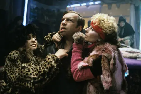 Scena z filmu Piotra Szulkina: mężczyzna w futrze w panterkę i różowym futrze, otoczony przez dwie kobiety w ekstrawaganckich strojach.