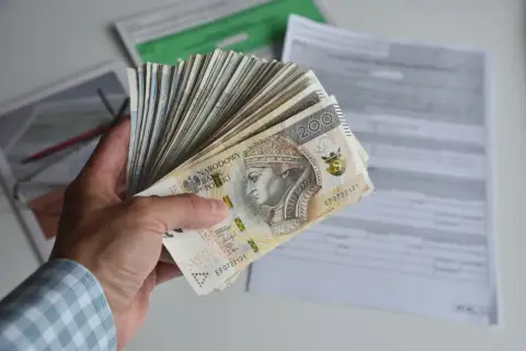 Dłoń trzyma plik banknotów 200 zł, symbolizujących dochód netto, na tle dokumentów podatkowych.