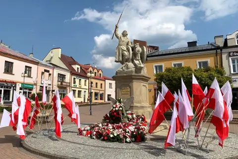 Pomnik z polskimi flagami, w tym kierunkowy 15, na placu przed kolorowymi kamienicami.