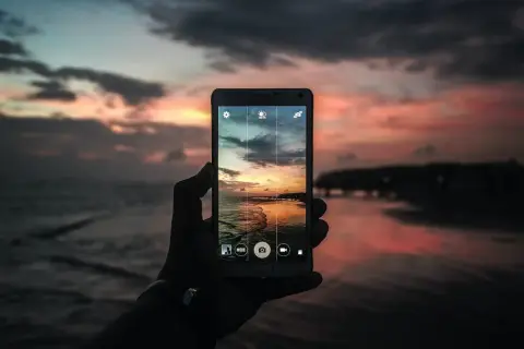 Mano che tiene un cellulare per scattare una foto con tramonto colorato sul mare.