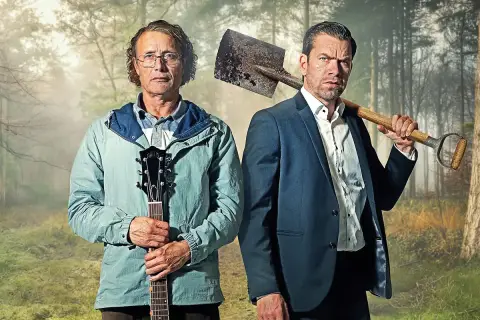 Två män i skogen, en med gitarr, en med spade. En scen från en mads mikkelsen serie.