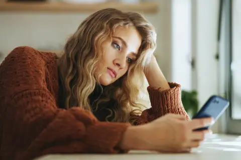 Młoda kobieta w brązowym swetrze, z ręką opartą o głowę, patrzy na telefon. Jej wyraz twarzy sugeruje anhedonię, brak radości z życia.