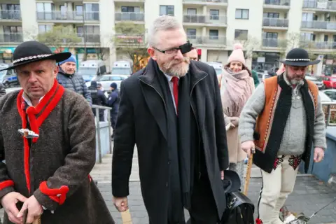 Grzegorz Braun w czarnym płaszczu i z opaską na oku, w otoczeniu mężczyzn w tradycyjnych strojach.
