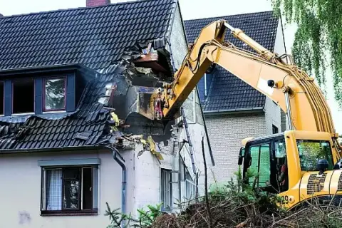 Żółta koparka prowadzi rozbiórkę dachu domu, niszcząc jego konstrukcję.