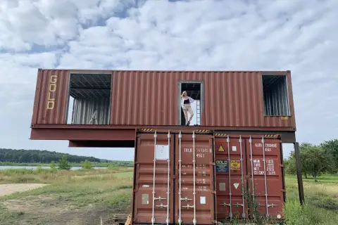 Kobieta stoi w drzwiach domu z kontenera morskiego, który jest częścią nietypowej budowli.