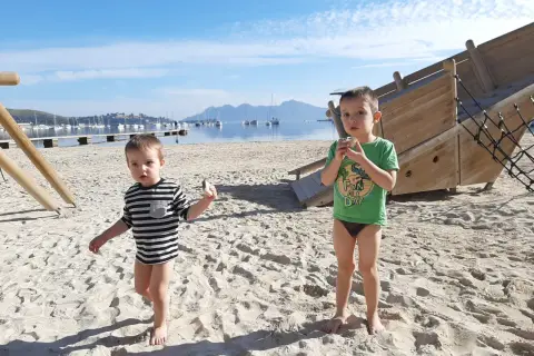 Dwóch chłopców na plaży, cieszących się słoneczną, majową pogodą w lutym. W tle widać morze z łodziami i góry.