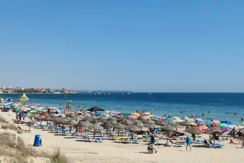 Día soleado en la playa de las Higuericas, con mucha gente disfrutando del mar y la arena bajo coloridas sombrillas.