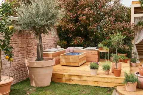 Ein idyllischer permakultur garten mit Olivenbaum, Zitruspflanze, Kräutern und Sitzgelegenheiten auf einer Holzterrasse.