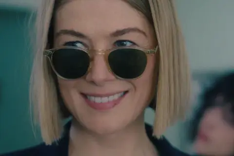 Uśmiechnięta Rosamund Pike w okularach przeciwsłonecznych, przypominająca scenę z jej filmów i seriali.