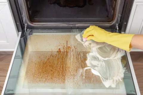 Ręka w żółtej rękawicy czyści zapieczoną szybę piekarnika. Czyszczenie piekarnika parowe to świetny sposób na pozbycie się trudnych zabrudzeń.