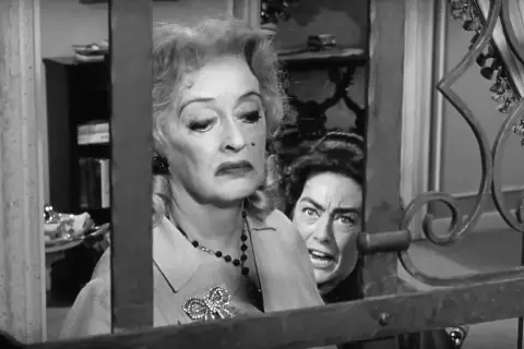 Bette Davis anziana, con un'espressione di disgusto, è dietro una grata. Un'altra donna con occhi sbarrati la spia.