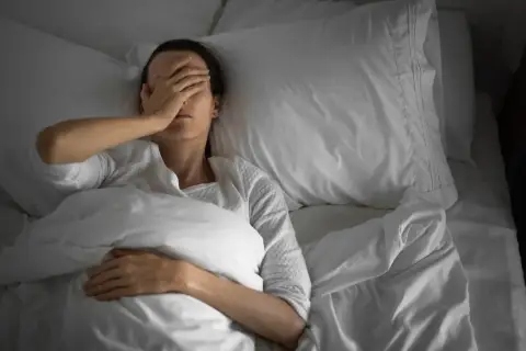 Frau liegt im Bett, bedeckt mit einer Decke, und hält sich die Hand vor die Augen. Sie kann nicht schlafen.