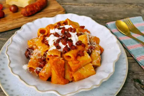 Un plato de macarrones con chorizo, cubiertos con queso rallado y una cucharada de crema. Al fondo, tomates y un trozo de queso.