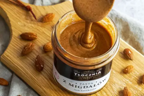 Gęsta, kremowa pasta z migdałów, czyli polskie "noisette", w słoiku, otoczona migdałami na drewnianej desce.