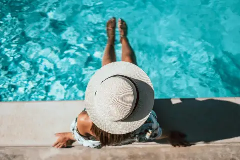 Kobieta w kapeluszu siedzi na brzegu basenu w ziemi, zanurzając nogi w turkusowej wodzie.