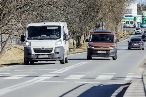 Biała furgonetka wyprzedza na przejściu dla pieszych pomarańczowego vana. W tle stacja benzynowa.