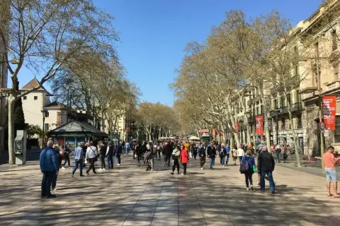 Tłum ludzi spaceruje po słonecznej La Rambla w Barcelonie, otoczonej drzewami i zabytkowymi budynkami.