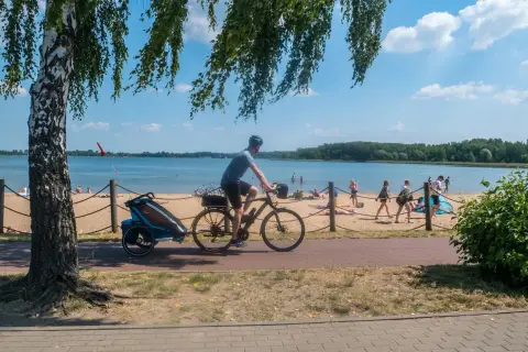Rowerzysta z przyczepką jedzie ścieżką przy plaży nad Pogorią. Ludzie odpoczywają na piasku.