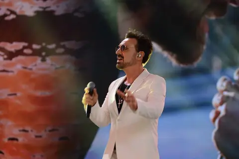 Mateusz Ziółko w białej marynarce śpiewa do mikrofonu na scenie.