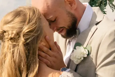 Agnieszka Kaczorowska i jej mąż w czułym uścisku podczas ślubu. Ona ma blond włosy splecione w warkocz, on w jasnej marynarce z butonierką.