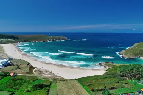 Vista aérea de la playa de Pantín con olas turquesas rompiendo en la arena dorada, rodeada de verdes colinas y el azul intenso del océano.