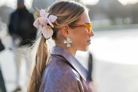 Mujer con coleta adornada con una flor de tela y pendientes de pétalos, luciendo gafas naranjas y una chaqueta de cuero.