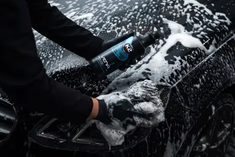 Ręce w czarnych rękawiczkach myją czarne auto pianą, trzymając butelkę szamponu.
