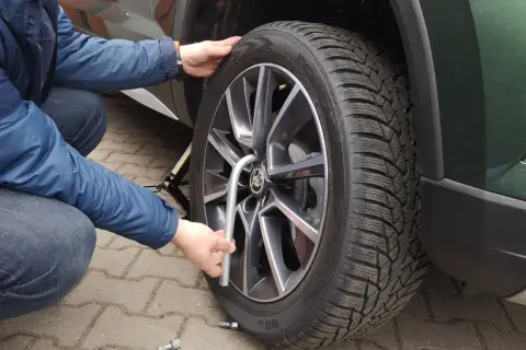 Osoba w niebieskiej kurtce dokręca śrubę koła samochodu kluczem.