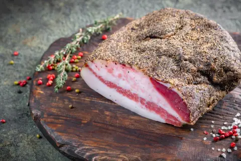 Guanciale co to? Włoski, peklowany policzek wieprzowy, pokryty pieprzem, leży na drewnianej desce z ziołami i pieprzem.