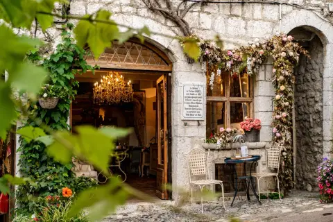 Urokliwa kamienna fasada w Saint-Paul-de-Vence, ozdobiona kwiatami i pnączami. W otwartych drzwiach wisi kryształowy żyrandol.