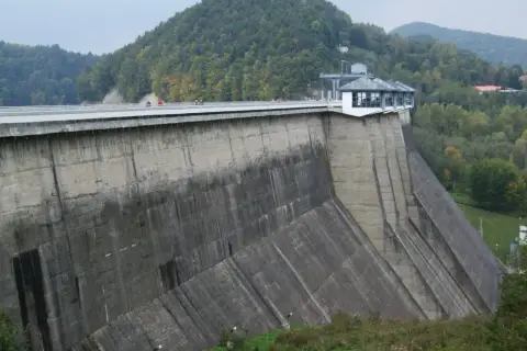 Ogromna zapora na sanie góruje nad zielonymi wzgórzami. Na jej szczycie widać ludzi spacerujących po moście.