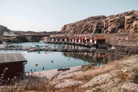 En rad röda sjöbodar längs en kuststig, med båtar i vattnet och en klippig bakgrund.