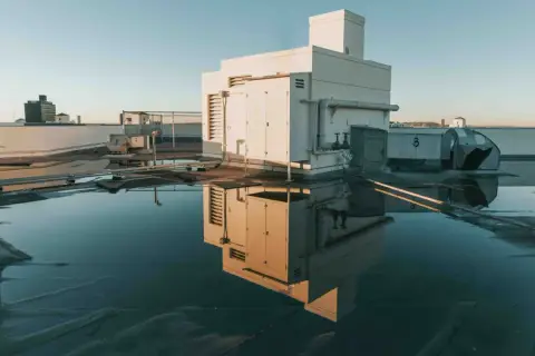 Flachdach mit Wasserpfützen, die ein Gebäude und Lüftungsanlagen spiegeln. Möglicherweise Anzeichen von Flachdach Blasenbildung unter der Oberfläche.