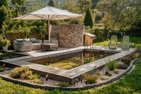 Ein schöner Garten mit einem teil-eingegrabenen Holzpool, Liegestühlen und einer gemütlichen Lounge-Ecke unter einem Sonnenschirm.