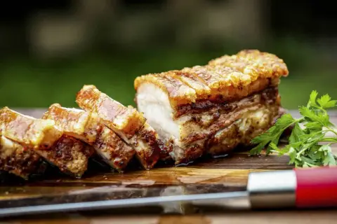 Ein perfekt gebratener, knuspriger Schweinebauch mit goldbrauner Kruste, auf einem Holzbrett mit Petersilie und einem Messer.