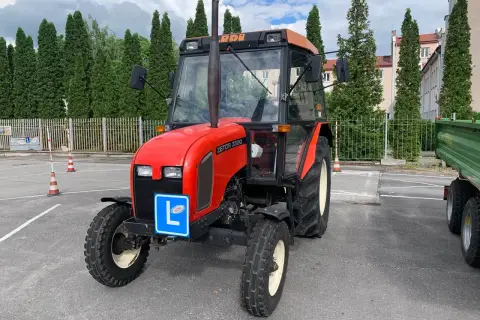 Czerwony traktor Zetor z literą "L" na niebieskiej tablicy, symbolizujący naukę jazdy. Kategoria t prawo jazdy.