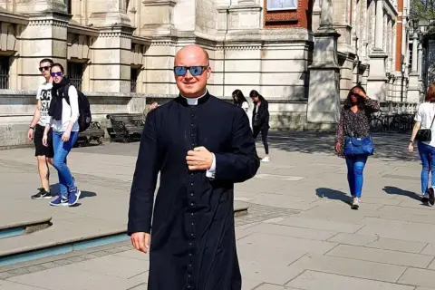 Uśmiechnięty pastor w czarnej sutannie i okularach przeciwsłonecznych idzie ulicą. W tle widać ludzi i budynek.