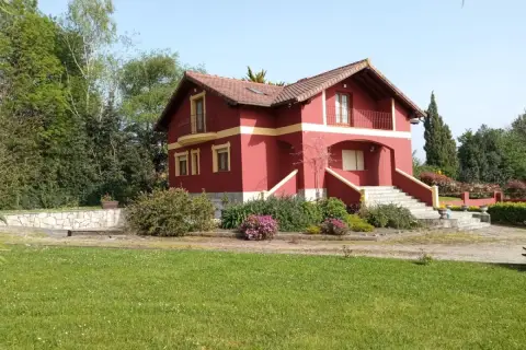 Una casa típica cántabra, de color rojo intenso con detalles amarillos, rodeada de vegetación y un césped verde.