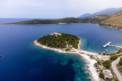 Isla con un castillo en Himare, Albania, rodeada de aguas azules y vegetación exuberante. Un muelle y barcos anclados completan la vista.