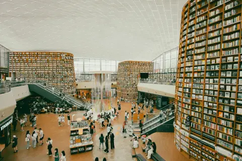 Wielopoziomowa biblioteka z półkami sięgającymi sufitu, gdzie każdy **bibliotekarz** marzyłby o pracy. Ludzie spacerują, czytają i odpoczywają wśród książek.