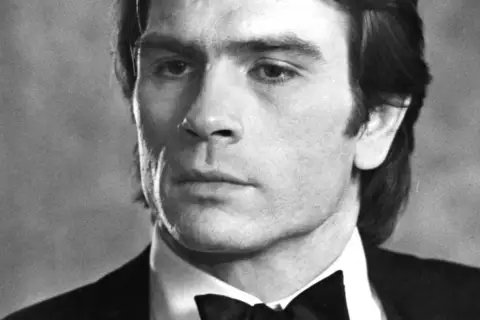 Młody Tommy Lee Jones w czarnym smokingu z muszką, zamyślony.